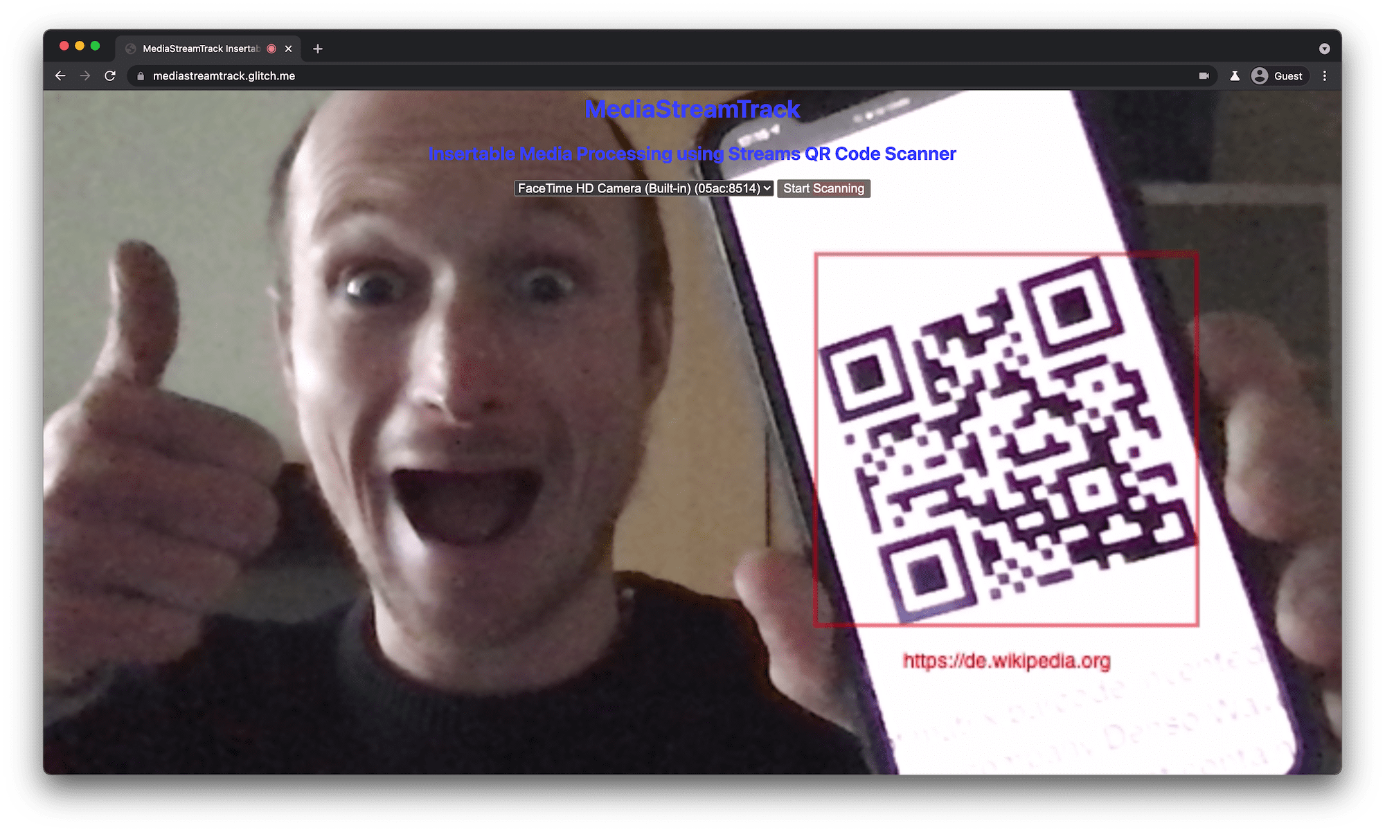 QR code scanner running in desktop browser tab showing a detected and highlighted QR code on the phone the user holds in front of the laptop's camera.